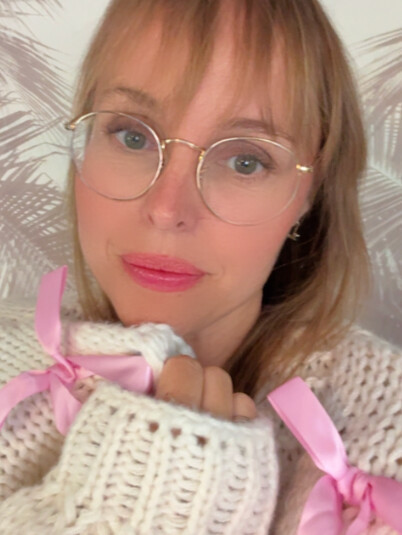 Portrait of Leah Bluewater in round glasses and a cream knit cardigan tied with pink ribbon bows, against a pale palm-leaf backdrop